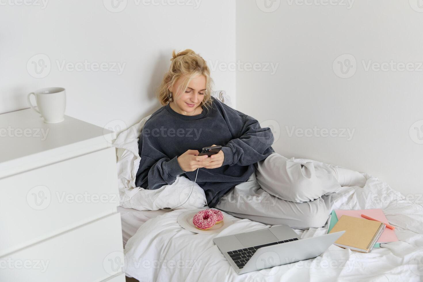 Lifestyle and people concept. Young beautiful woman, staying at home, lying in bed with laptop and smartphone, eating doughnut, enjoying free time, spending weekend at home, watching movie online photo