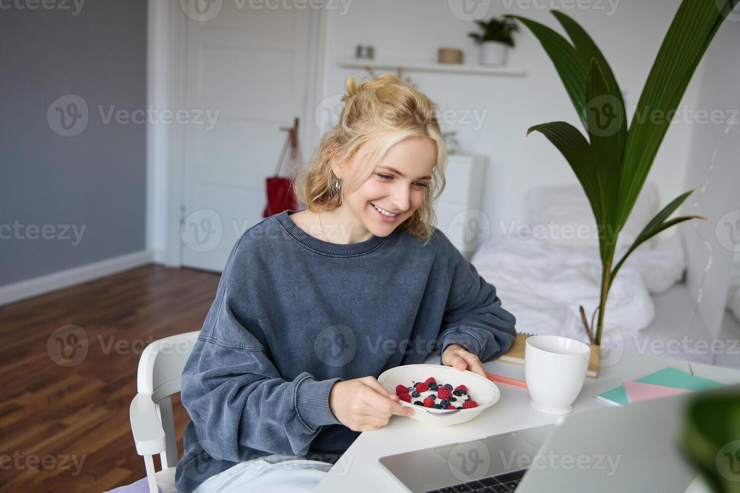 retrato de joven sincero niña acecho s en computadora portátil, disfrutando película y comiendo en frente de computadora portátil, teniendo desayuno y curioso a pantalla foto
