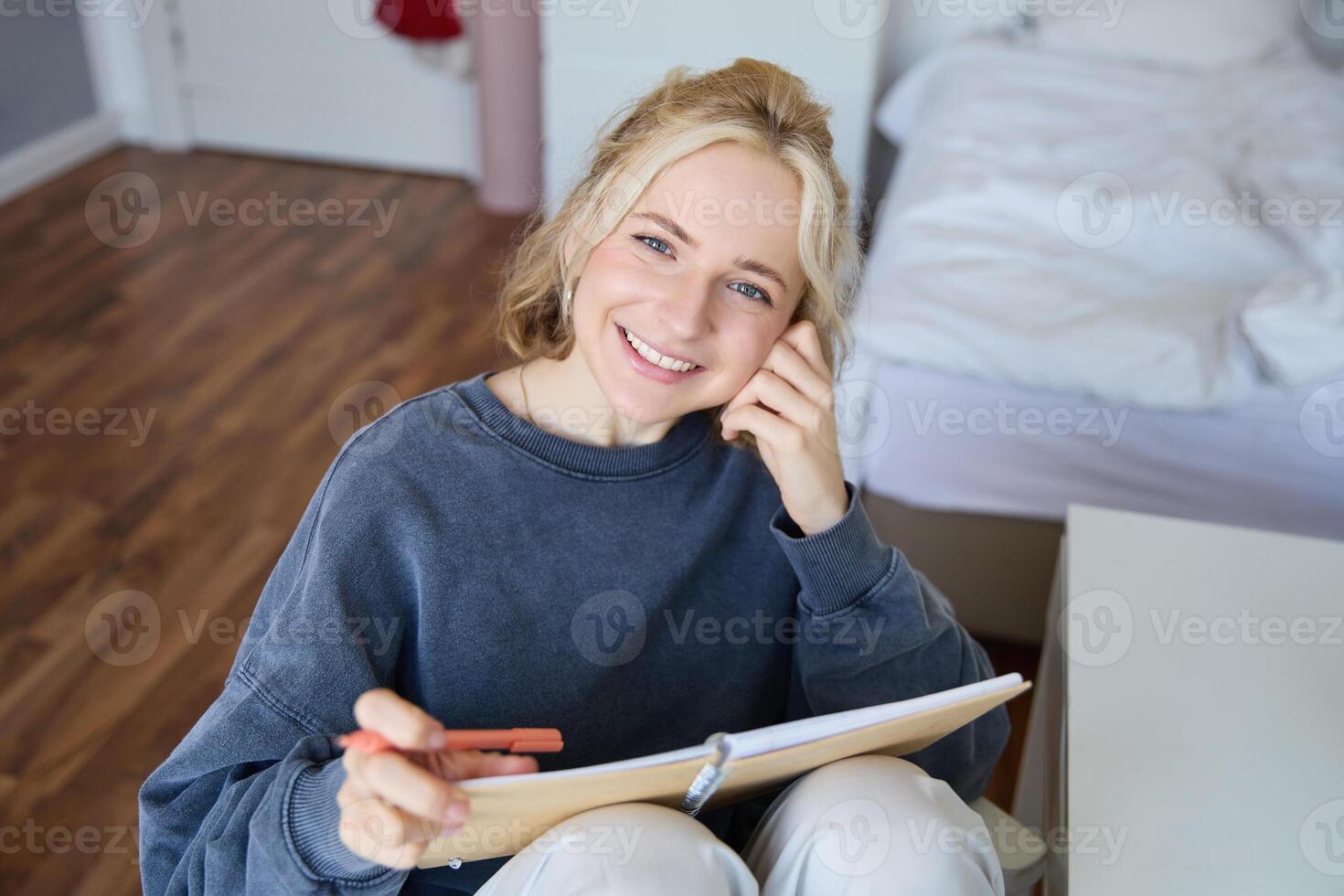 Portrait of smiling, beautiful young candid woman, sitting in bedroom with pen and notebook, writing in journal, making notes, looking happy at camera photo