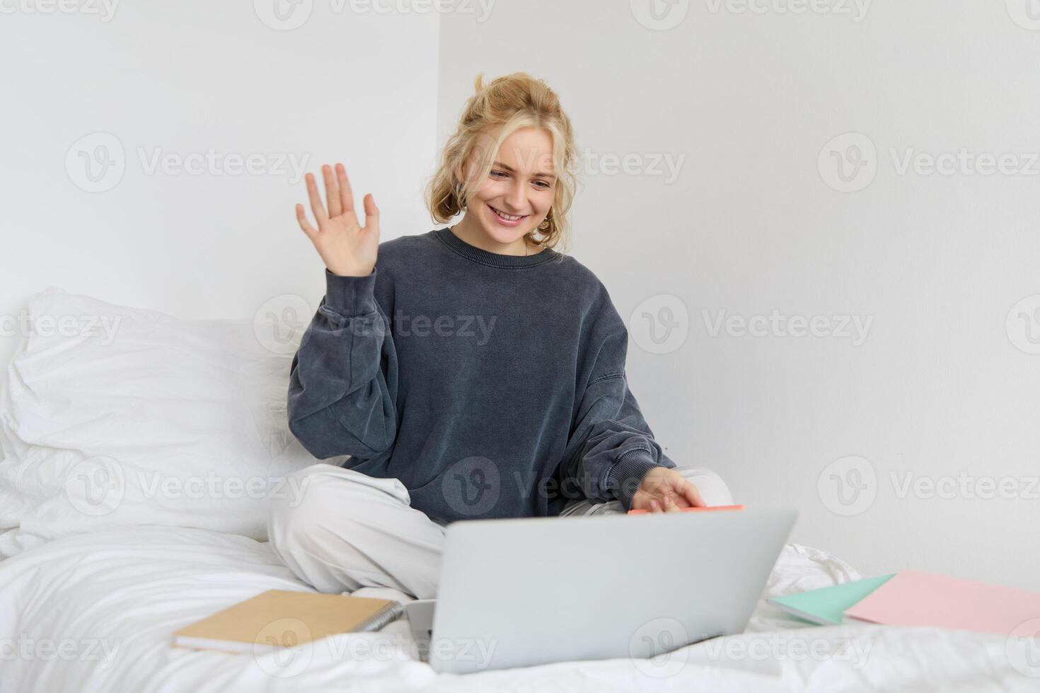 Portrait of young happy woman, connects to chat, using laptop, waving hand at camera, saying hello to someone online, sitting on bed, studying, e-learning or working from home photo