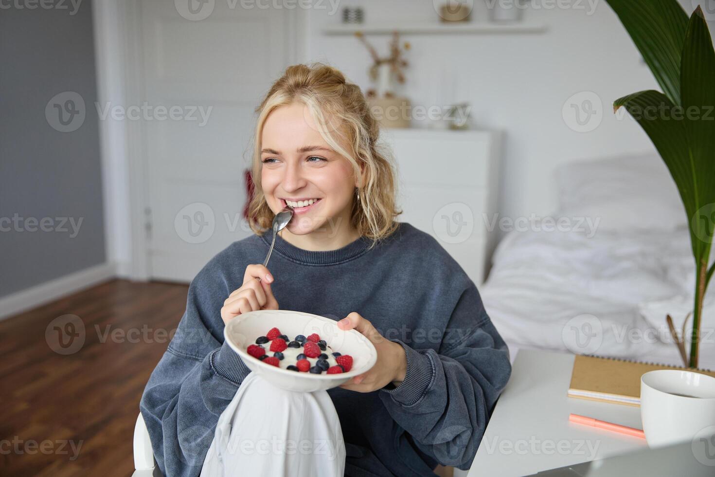 Close up portrait of smiling, cute blond woman, eating healthy lunch in her room, holding bowl with berries, biting spoon, looking happy photo