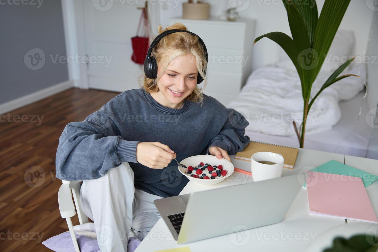Portrait of happy young blond woman, sitting in a room, watching movie on laptop and eating healthy breakfast, drinking tea, resting on weekend photo