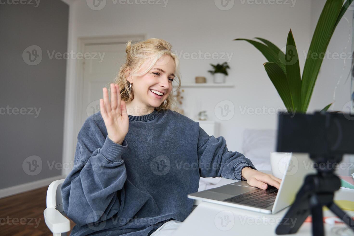 Portrait of young social media content creator, woman sits in room with laptop and digital camera, records lifestyle vlog, says hello, chats to someone online via computer photo