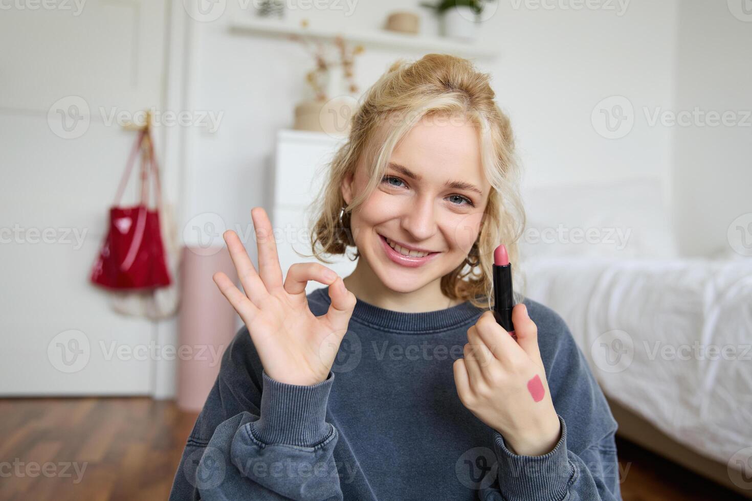 Portrait of young blond woman, content creator, recording for social media about makeup, showing okay hand sign and lipstick, recommending good cosmetic product photo