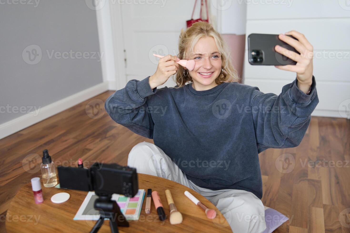 Image of stylish young woman, social media influencer, taking pictures on mobile phone, doing makeup tutorial for followers online, recording vlog in her bedroom, showing brush photo