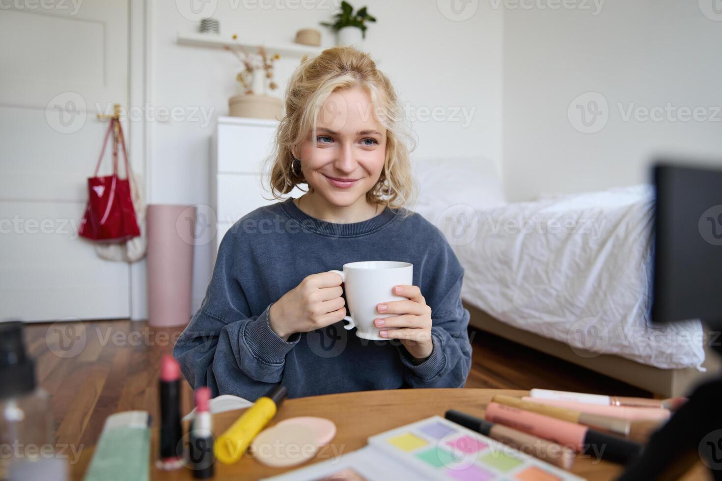 Image of young woman, makeup vlogger, sitting in bedroom with digital camera, drinking tea and talking, creating lifestyle , social media content photo