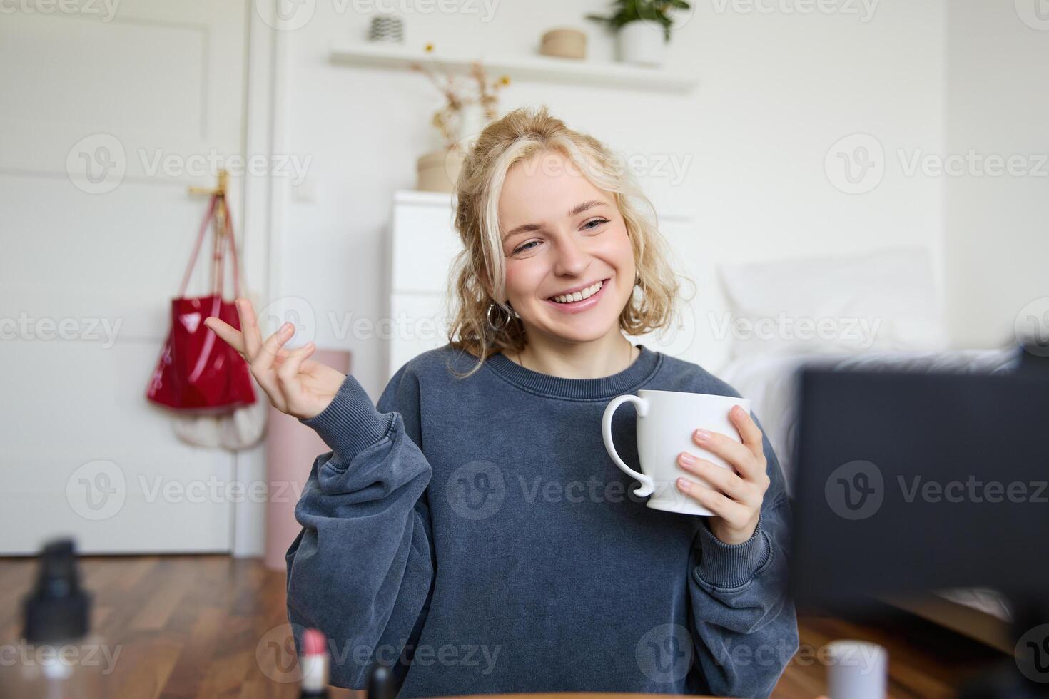 Portrait of cute smiling woman, blond girl drinks tea and records a casual, lifestyle blog, vlogger sits in a room with camera and stabiliser, holds cup of coffee and talks photo