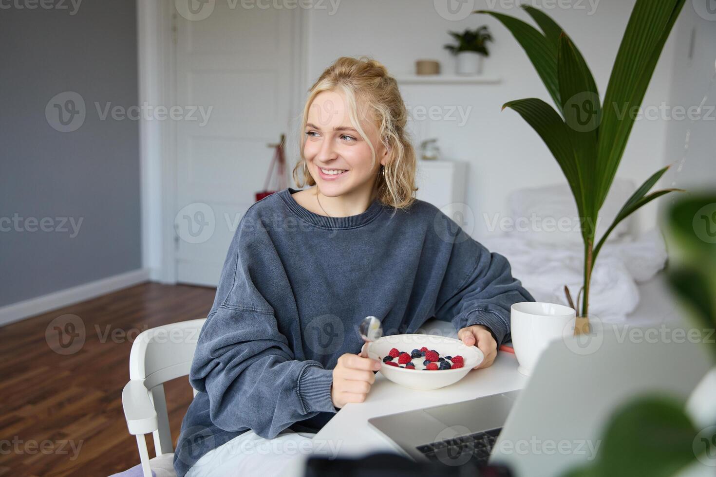 Portrait of young candid girl watching s on laptop, enjoying movie and eating in front of laptop, having breakfast and staring at screen photo
