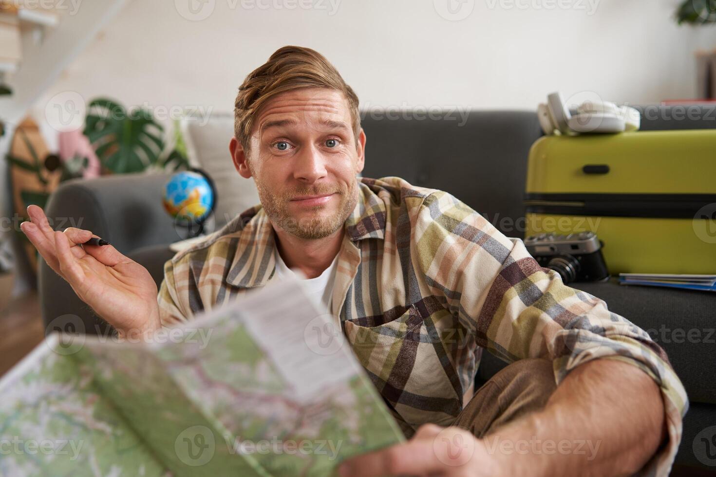 Image of shrugging man with road travel map, looks at camera with confused face, packs suitcase for holiday, plans a trip photo