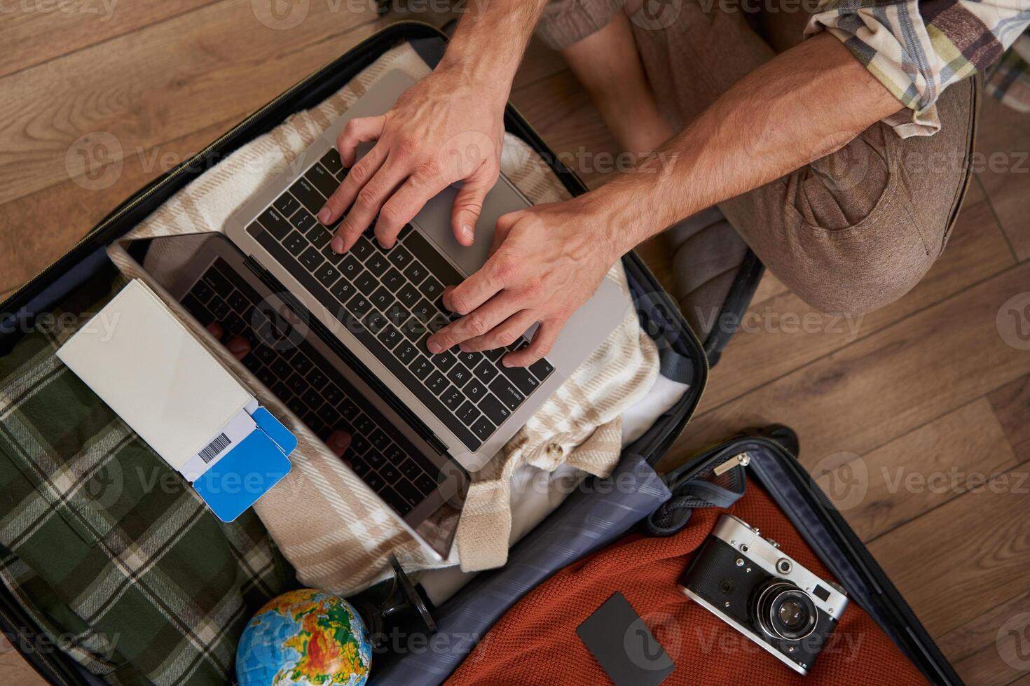 Top view of opened suitcase with clothes, male hands using laptop, booking hotel for vacation, check-in on flight for holiday, packed camera to shoot pictures photo