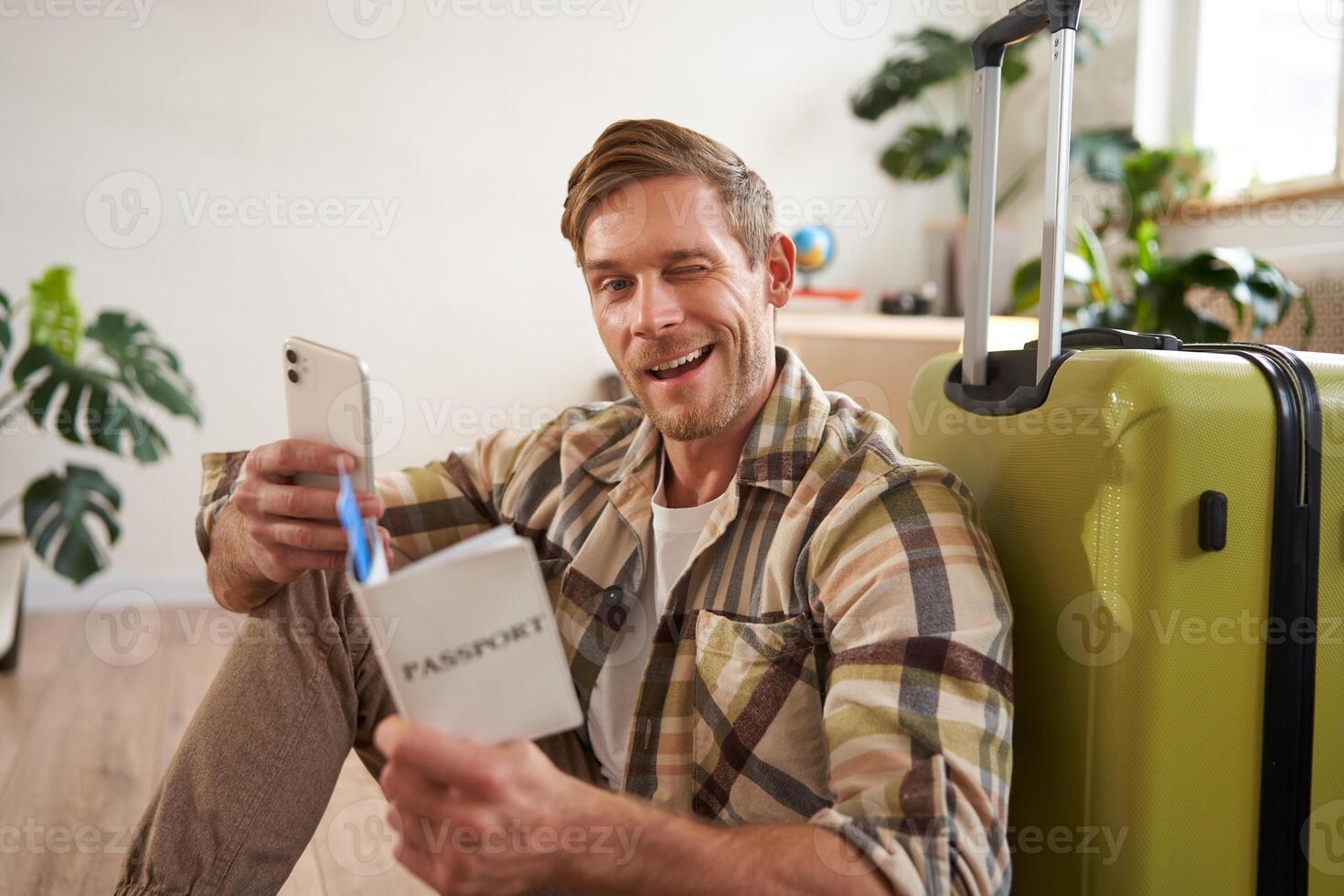 Charismatic young man, traveller going on holiday, trip abroad, sitting with suitcase, holding smartphone, tickets and passport, winking and smiling at camera photo