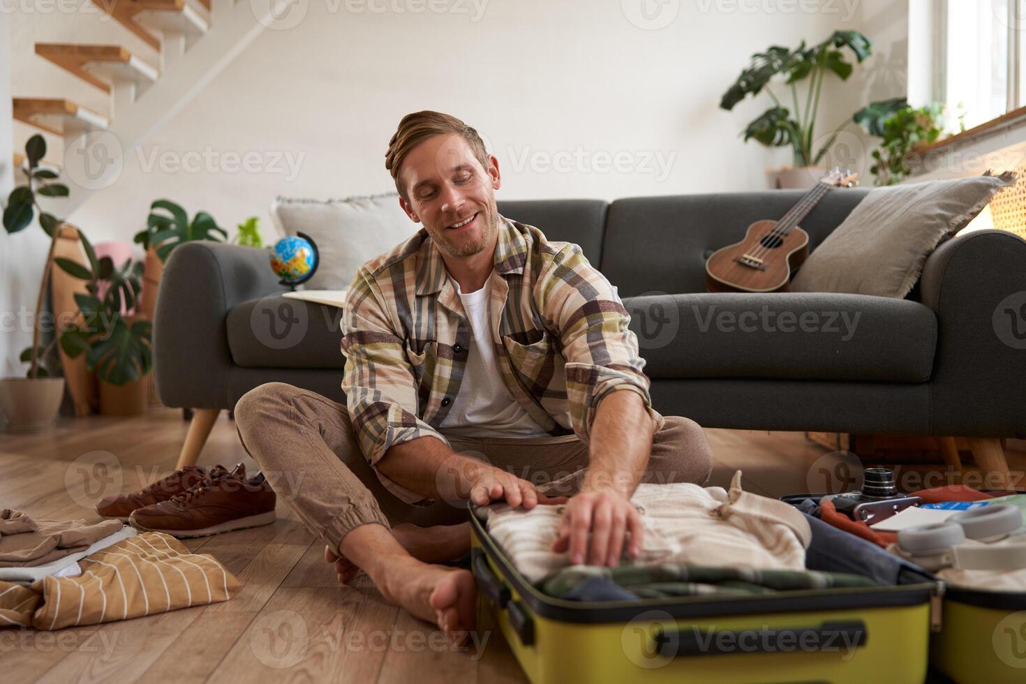 Image of young man, traveller, sitting on floor with opened suitcase, packing clothes for a trip, going on holiday photo