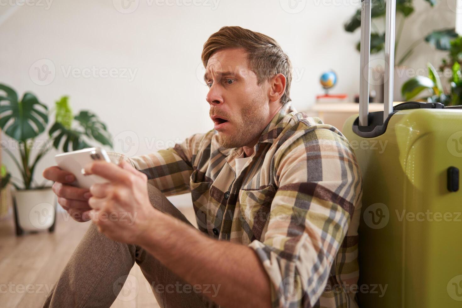 Man with shocked face, sitting on floor with suitcase, looking worried and tensed on mobile phone screen photo