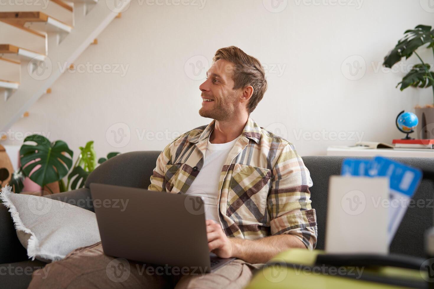 Portrait of handsome young man, has two tickets and passport, goes online on laptop, booking hotel and planning his holiday, sitting in living room photo