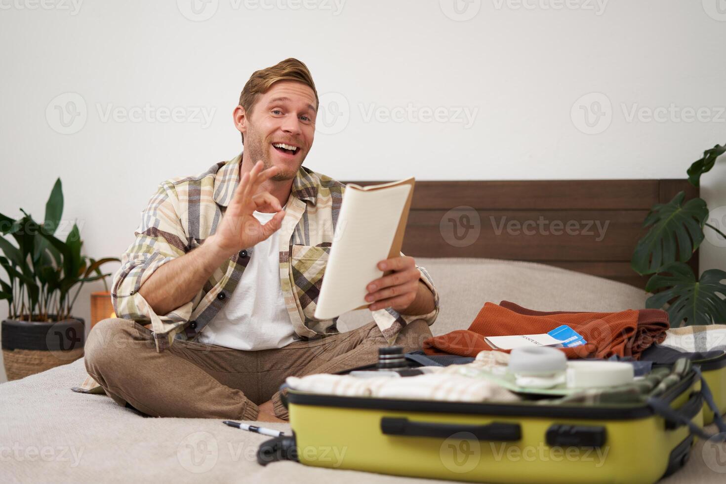 Image of cheerful man shows okay sign, packs clothes in suitcase, goes on holiday or business trip, holds notebook with list of items to take with him photo
