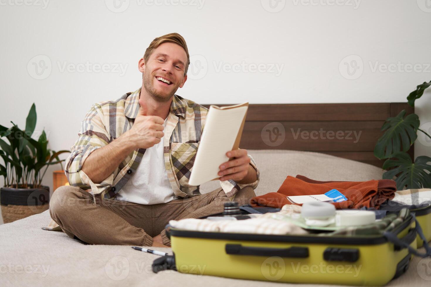 alegre joven hombre, turista con cheque lista, demostración su notas en computadora portátil, pulgares arriba, embalaje maleta, yendo en día festivo, de viaje extranjero y preparando equipaje foto