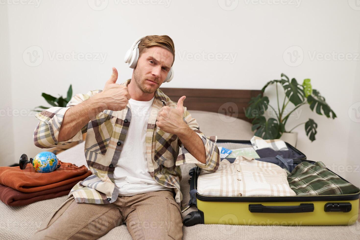 Portrait of cheerful handsome man, tourist going on vacation, dancing and listening to music in headphones while packing suitcase with clothes for holiday trip abroad photo