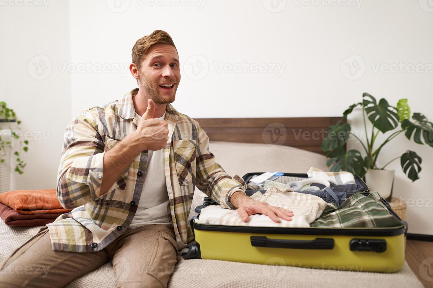 Attractive young man smiling, showing thumbs up, showing his clothes inside suitcase, getting ready for a holiday, travelling with travel agency tour photo