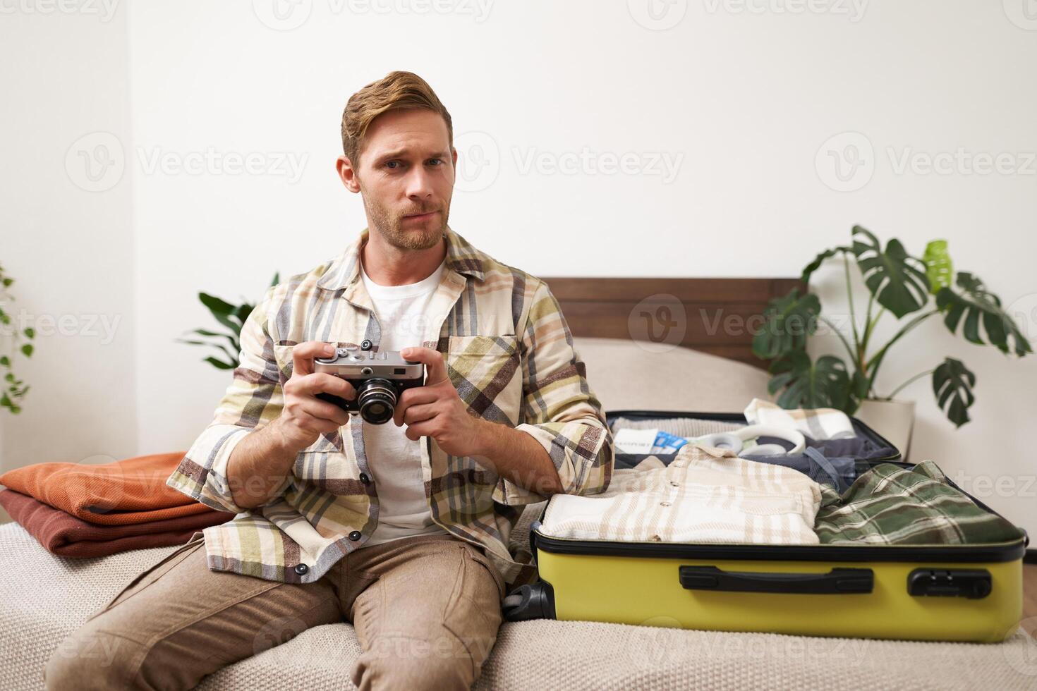 Portrait of handsome man going on vacation, packing suitcase, holding camera, travel photographer going on holiday photo