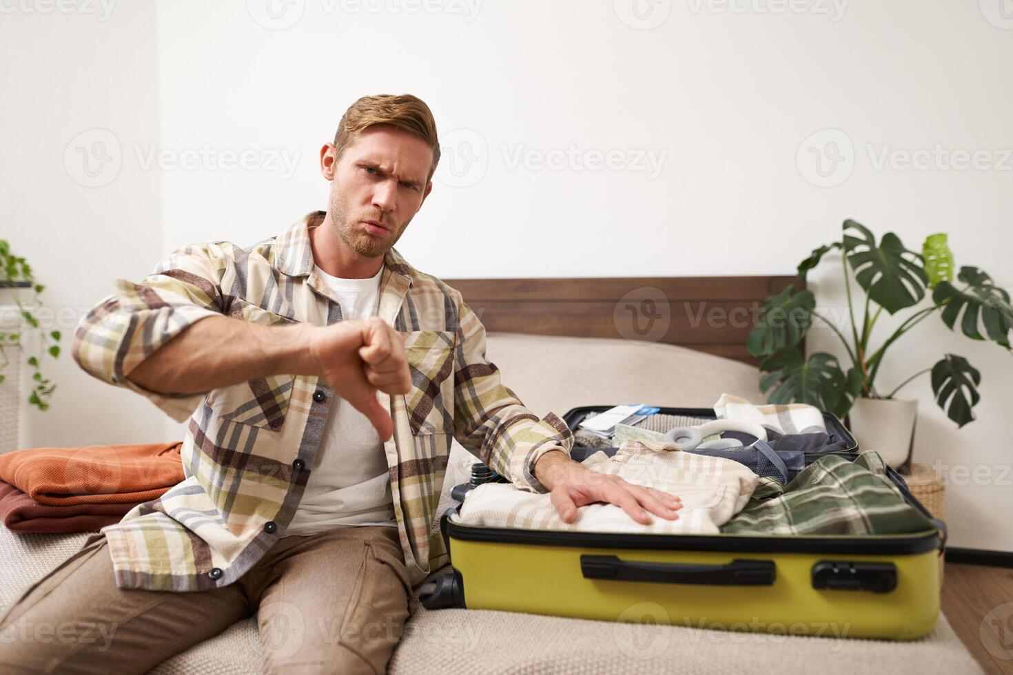 Image of young disappointed man, showing thumbs down, puts hand on his clothes inside luggage, dislike, disapprove something awful, upset with his holiday photo