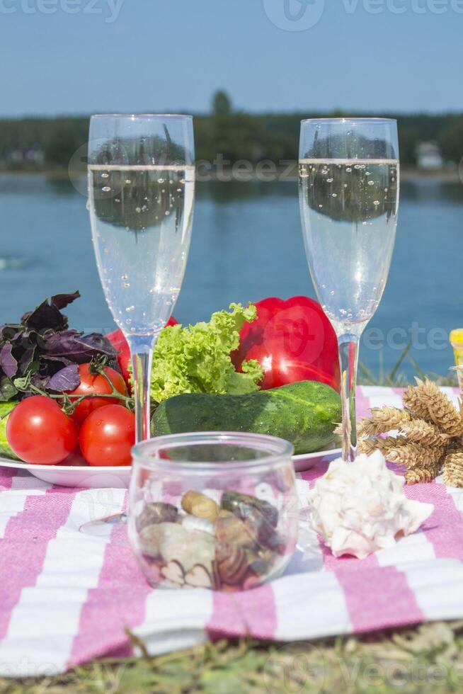 Beautiful picnic with fruits, vegetables and glasses of wine near lake. photo