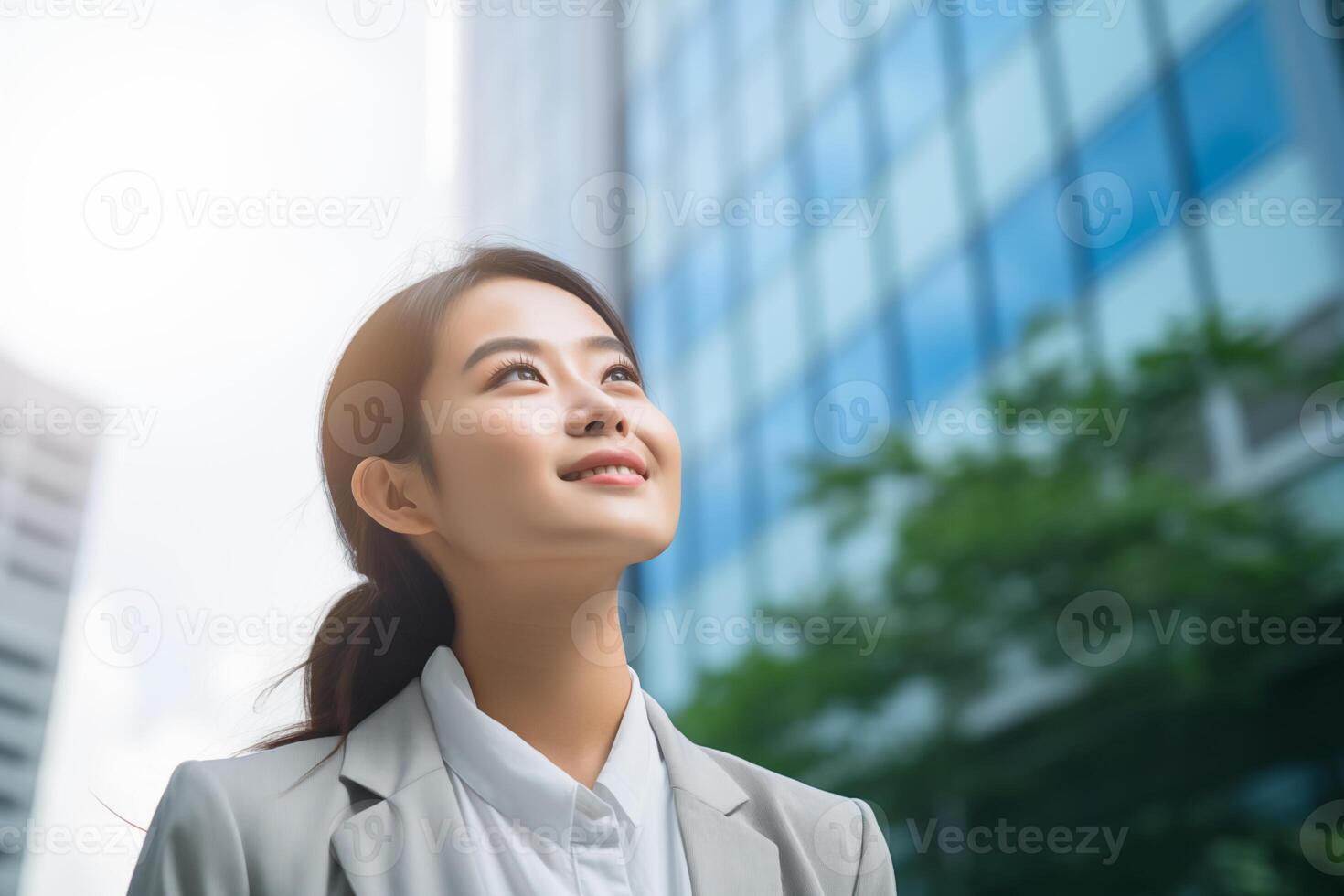 contento joven asiático negocio mujer en pie en calle fuera de moderno oficina edificio en el ciudad. retrato de profesional mujer de negocios. oficina trabajador mirando arriba a el cielo y sonriente foto