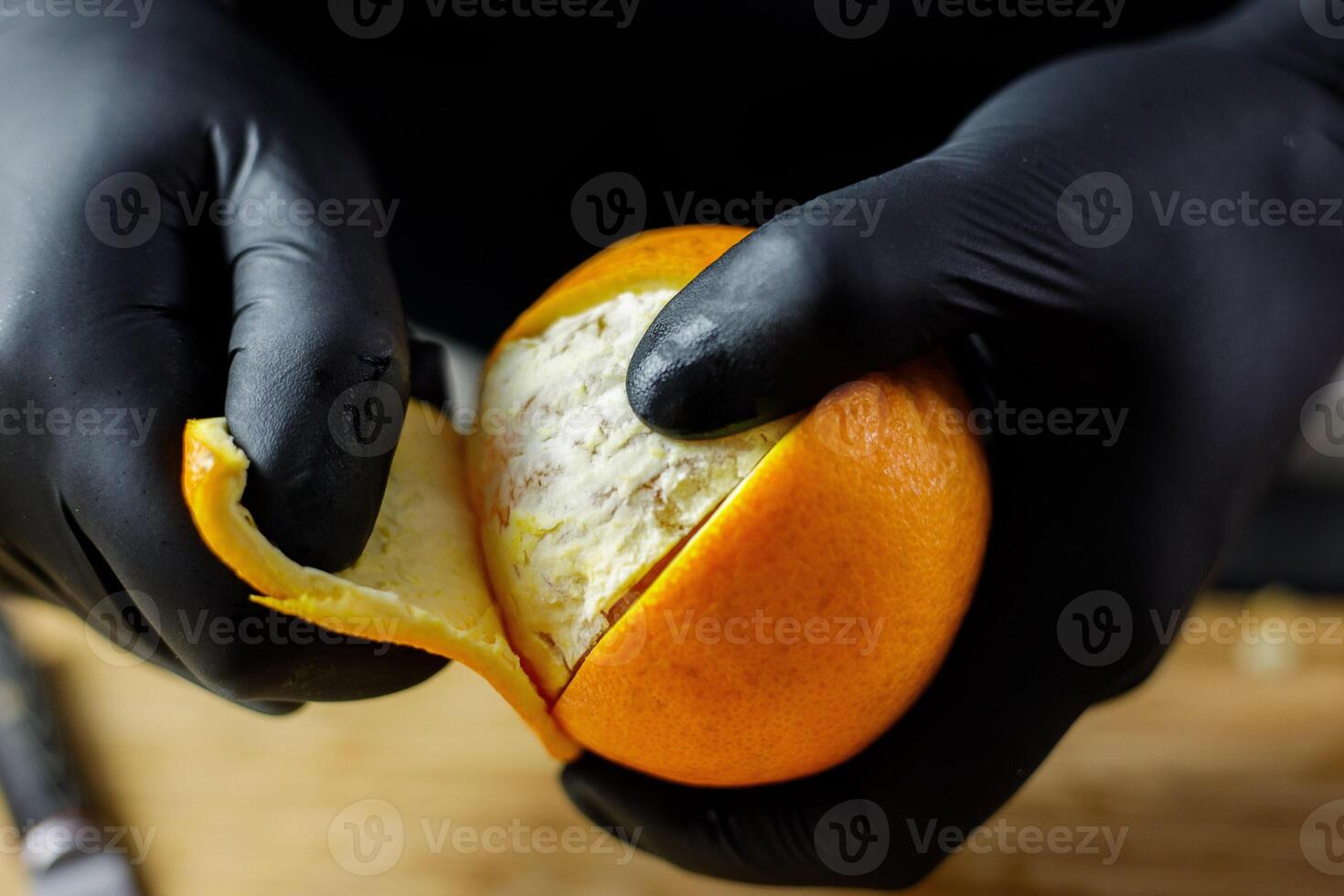 Person with black gloves peeling full orange on wooden cutting board photo