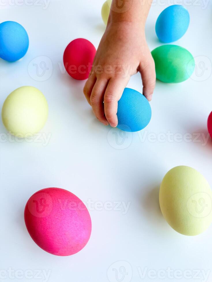 Childs hand picking up a blue Easter egg among colorful eggs on a white surface, interactive and engaging holiday activity concept. Can be used for educational content highlighting fine motor skills photo