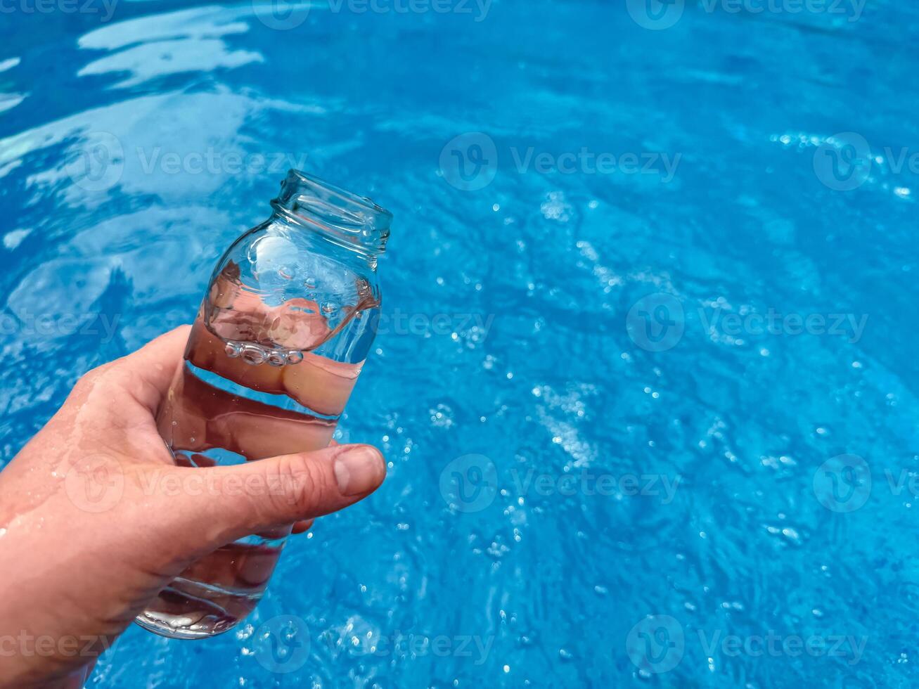 Hand holding clear glass bottle partially filled with water against sparkling blue water background with empty space for text. Clean water concept. photo