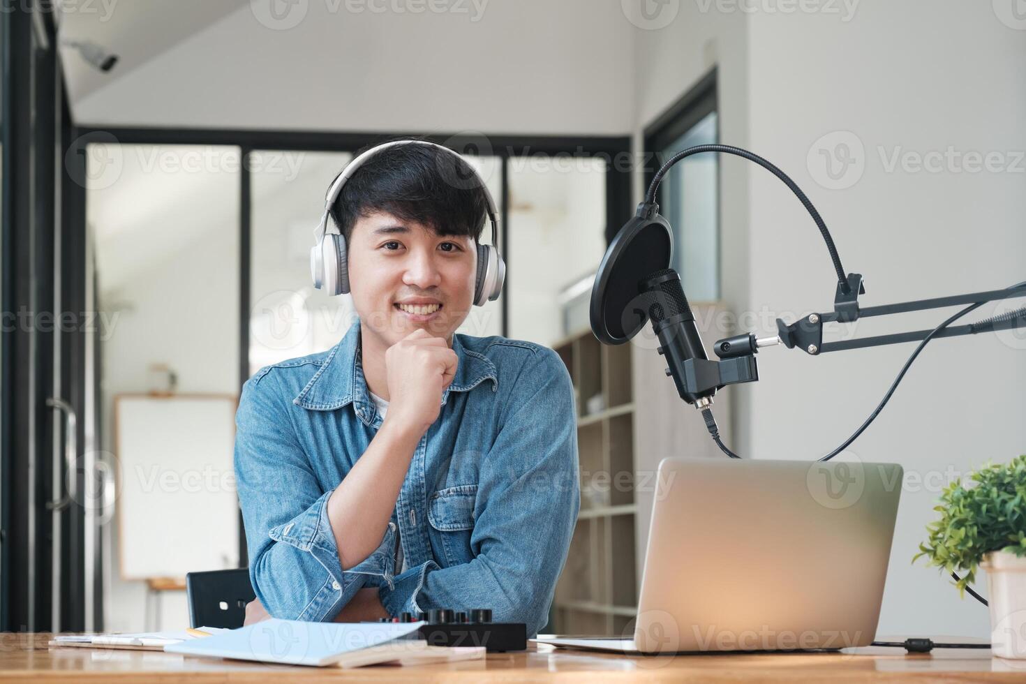 A man wearing headphones is sitting at a desk with a laptop and a notebook photo