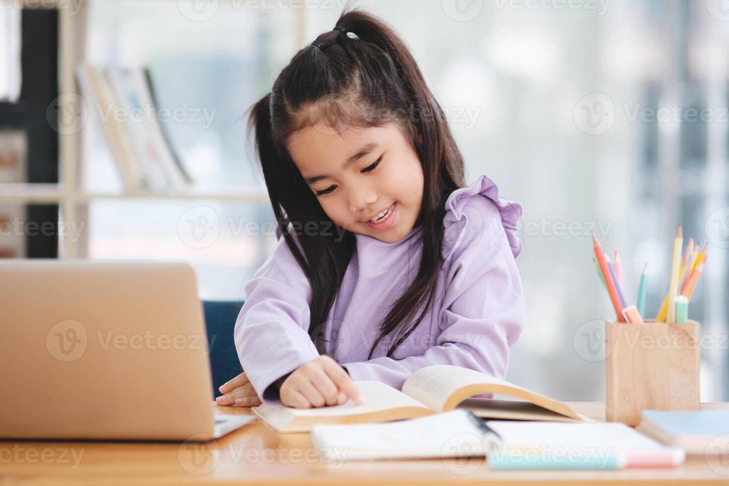 A young girl is sitting at a desk with a laptop and a book photo