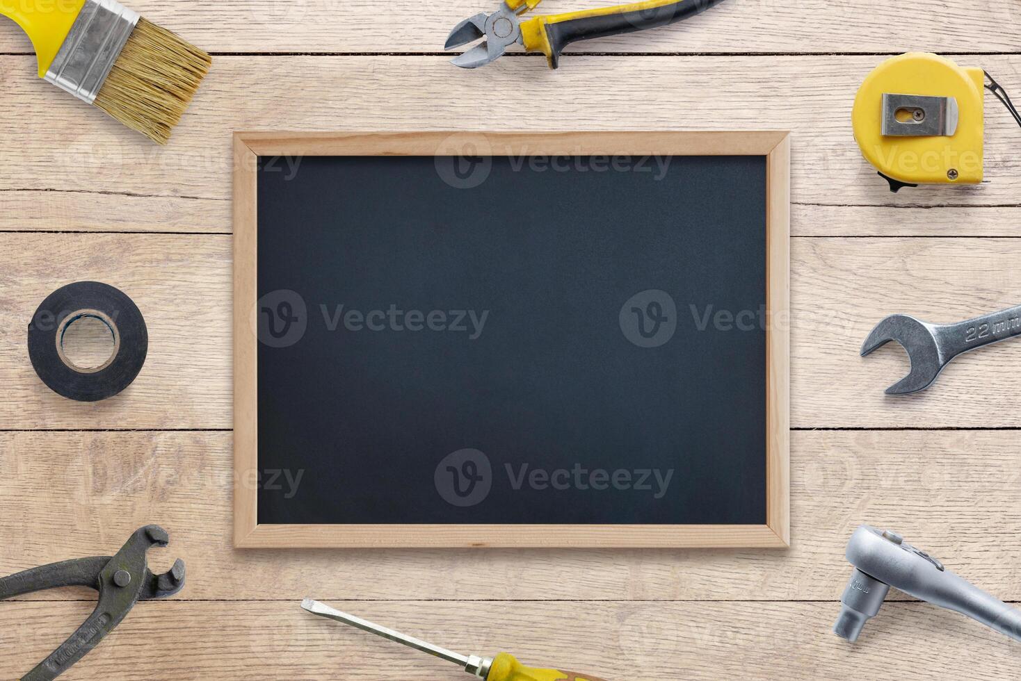 Clean chalkboard surrounded by tools, brush, pliers, screwdriver, tape measure, adhesive tape, wrench on wooden table. Perfect for text, logo promotion. Top view, flat lay composition. DIY concept photo