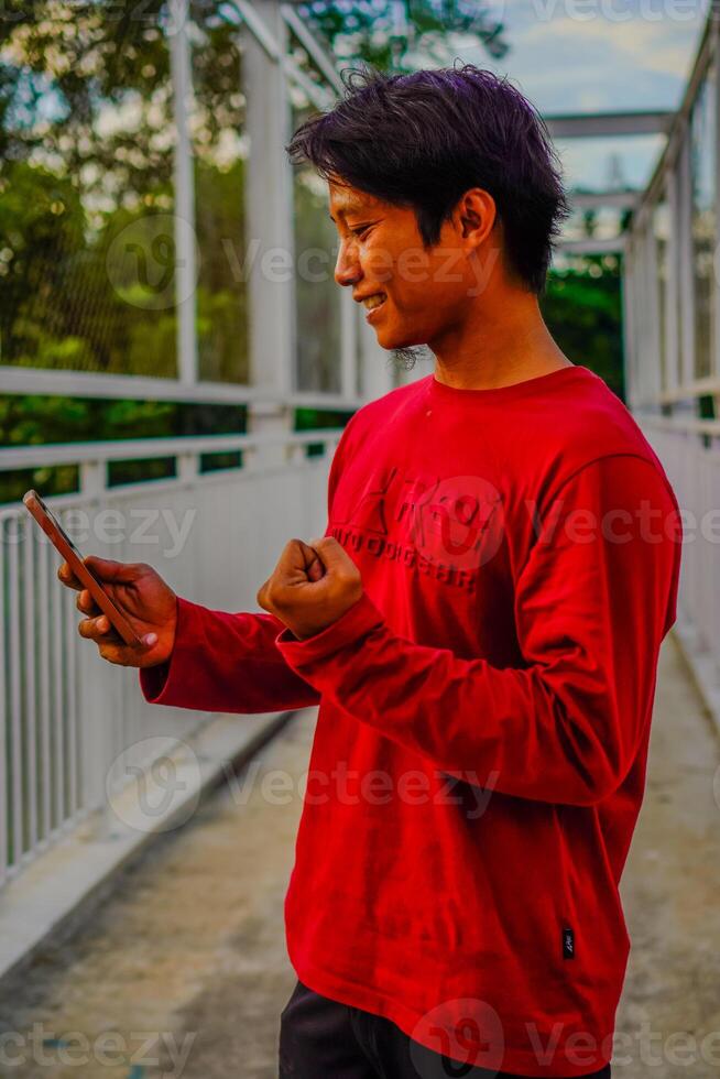 javanés hombre en rojo es mirando a su Teléfono móvil con un contento expresión. foto