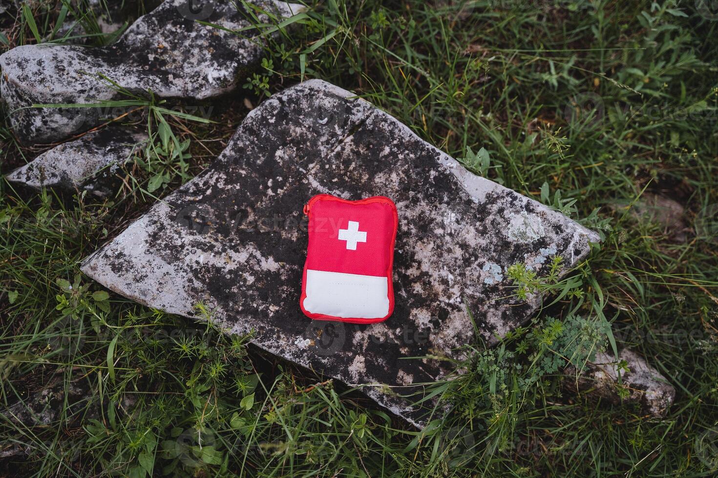 A red first aid kit lies on a stone in nature, a small hiking bag with essential medicines, a white cross symbol, a medical bag. photo