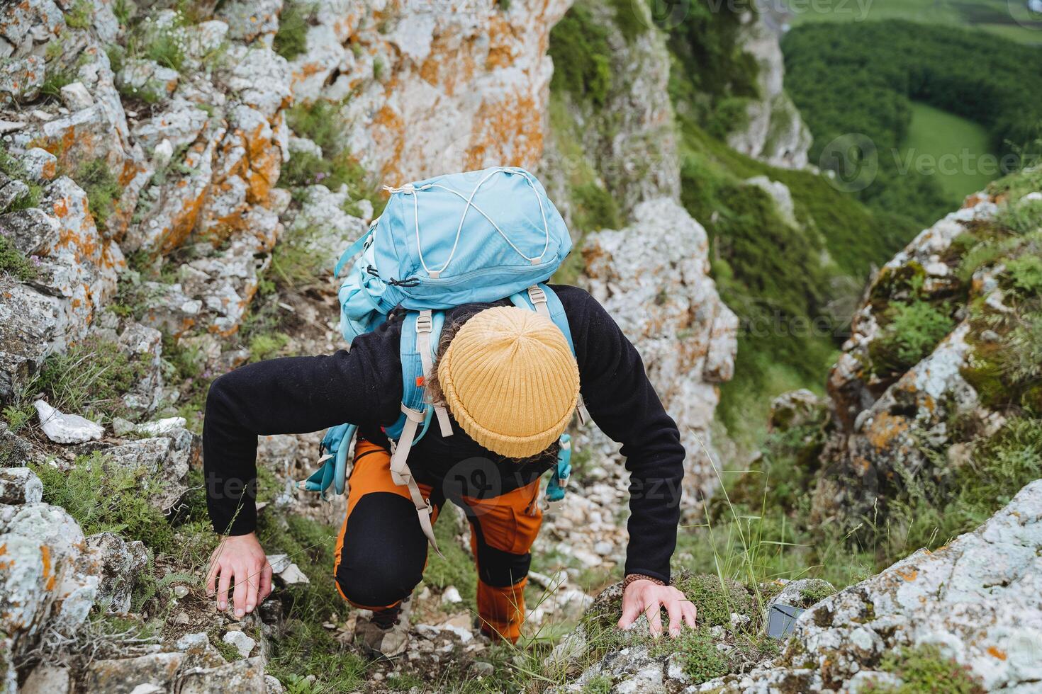 A tourist climbs into the mountains clinging to stones with his hands, a heavy backpack of a tourist on his back, a yellow warm hat, climbing, a man in the mountains. photo