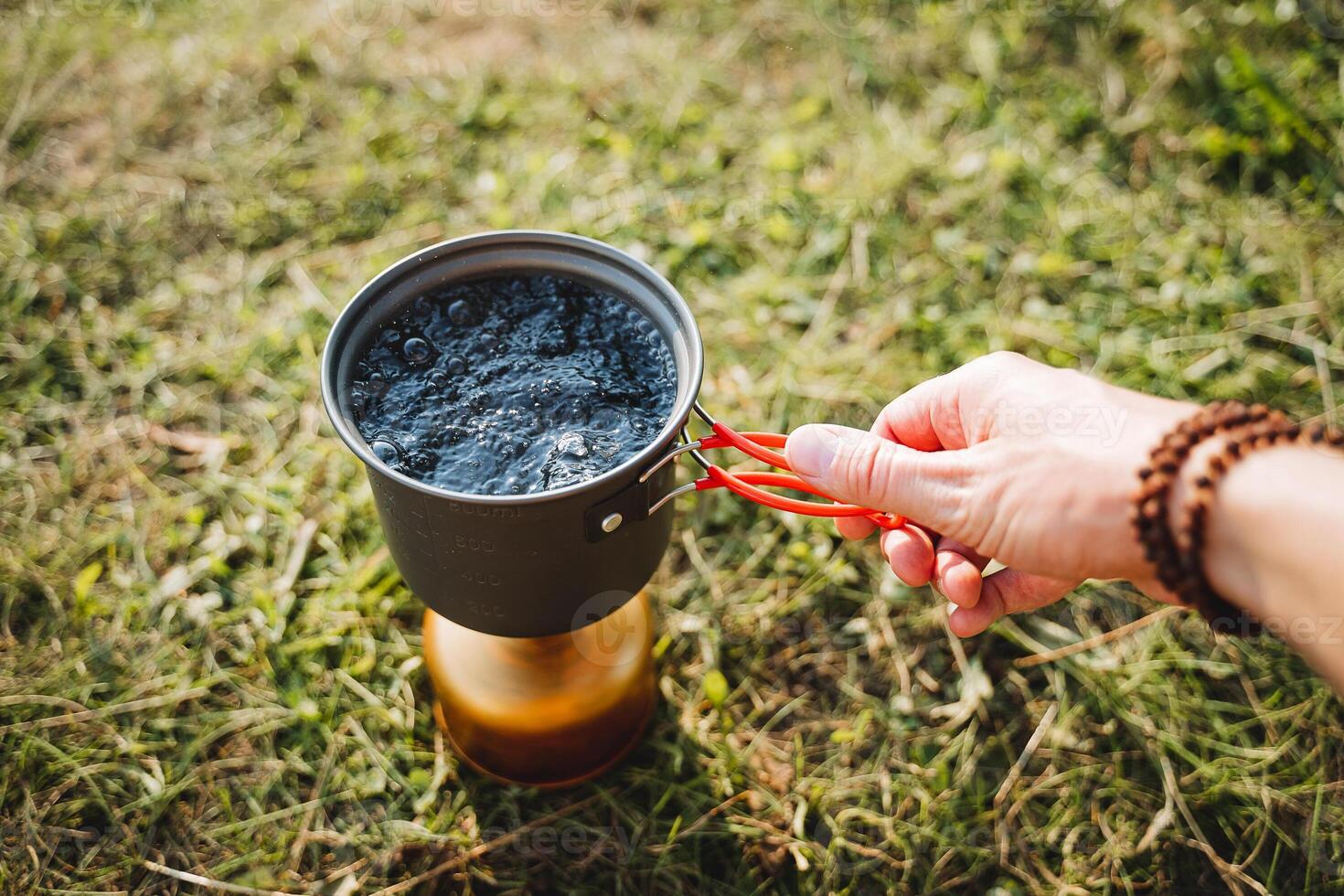 Hand holds a pot of hot water, a tourist cooks food on a hike on a gas burner, tourist utensils, camping in the forest, boil water for tea. photo