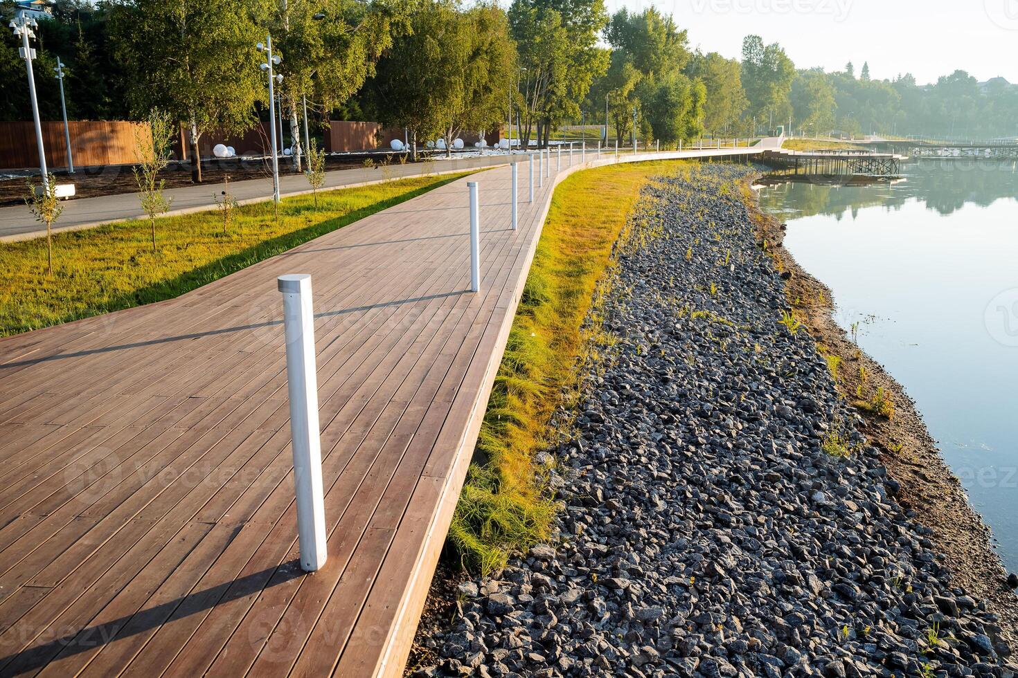 A new embankment in the city park near the water, a beautiful landscape design of the park, a wooden road for pedestrians, lampposts, a stone bank. photo