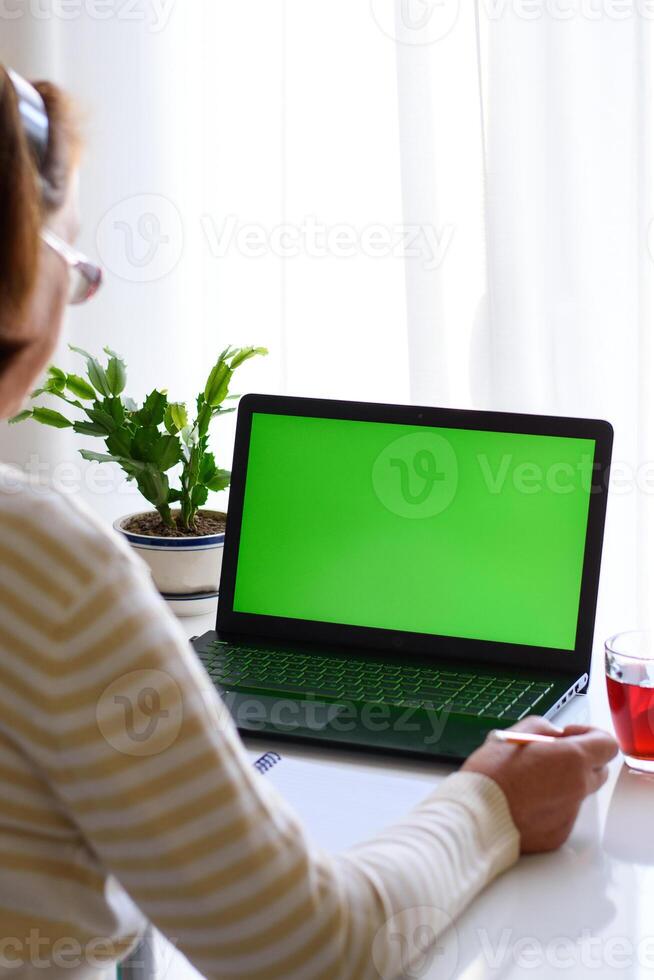 Vertical back over shoulder view of elderly woman with glasses watching educational webinar, online lecture seminar on laptop with green screen, taking notes. The concept of distance education. photo