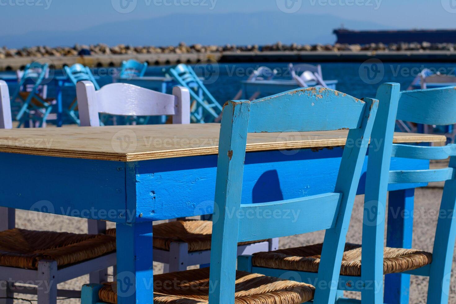 Closeup on wooden table and chair in typical fish tavern at fishing port. Selective focus. Big ship on background. Aegean island Chios in Greece on an autumn day. Greek holidays and destinations photo