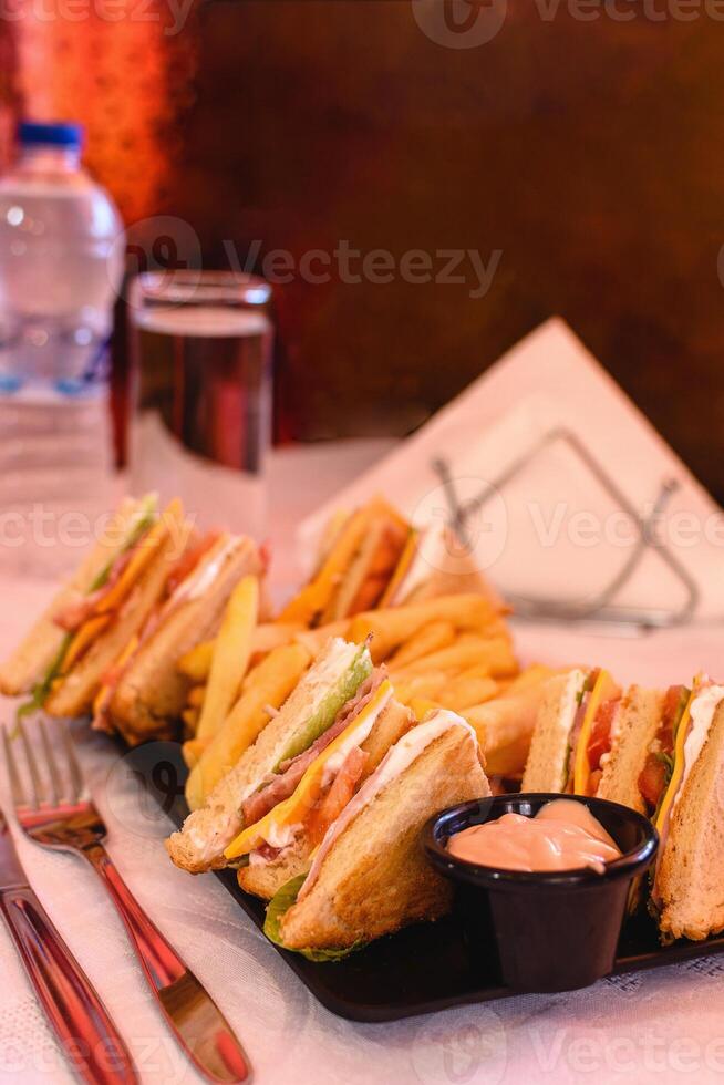 Vertical view of classic club sandwich with ham, cheese, bacon, tomato, lettuce, mayonnaise served with fries on the black plate on white tablecloth in greek tavern. Table setting. Soft focus photo