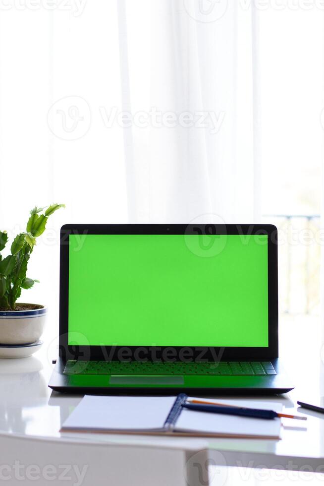 desk setups, work environments. Vertical view of laptop with green screen, notebook, pen and flower on the table in front of window. Selective focus. Concept of distance education photo
