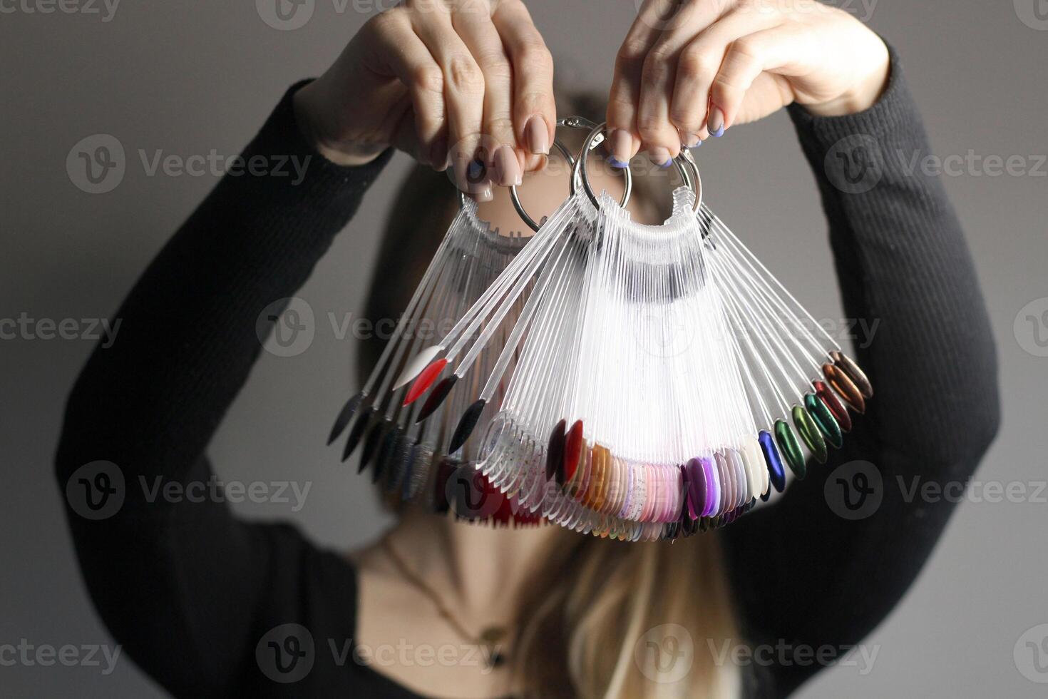 el niña sostiene en su manos un paleta de muestras de de colores uña pulimentos el cara es no visible. gris antecedentes. uña color polaco mostrar. escoger el color de uña polaco foto