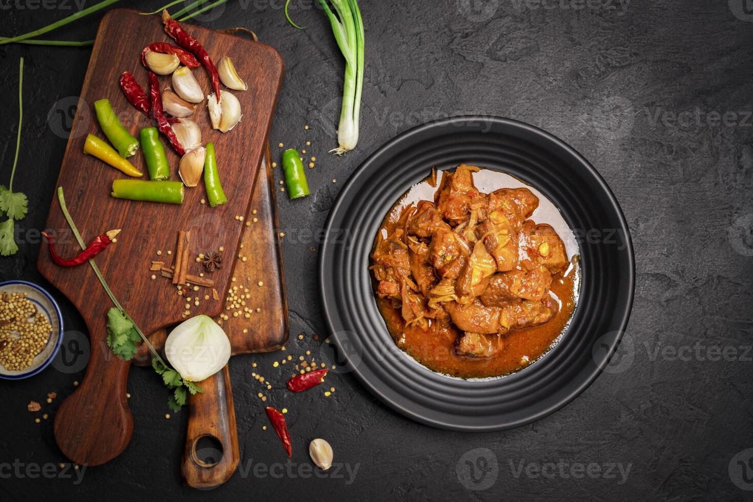 Delicious beef or pork stew in a rustic black plate with fresh coriander, red chilies, garlic, onions and spices placed on a black background, top view photo