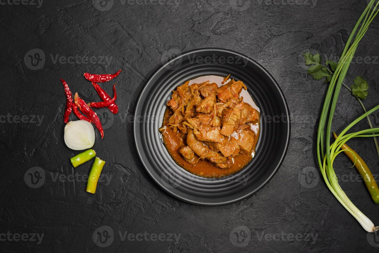 Delicious beef or pork stew in a rustic black plate with fresh coriander, red chilies, garlic, onions and spices placed on a black background, top view photo