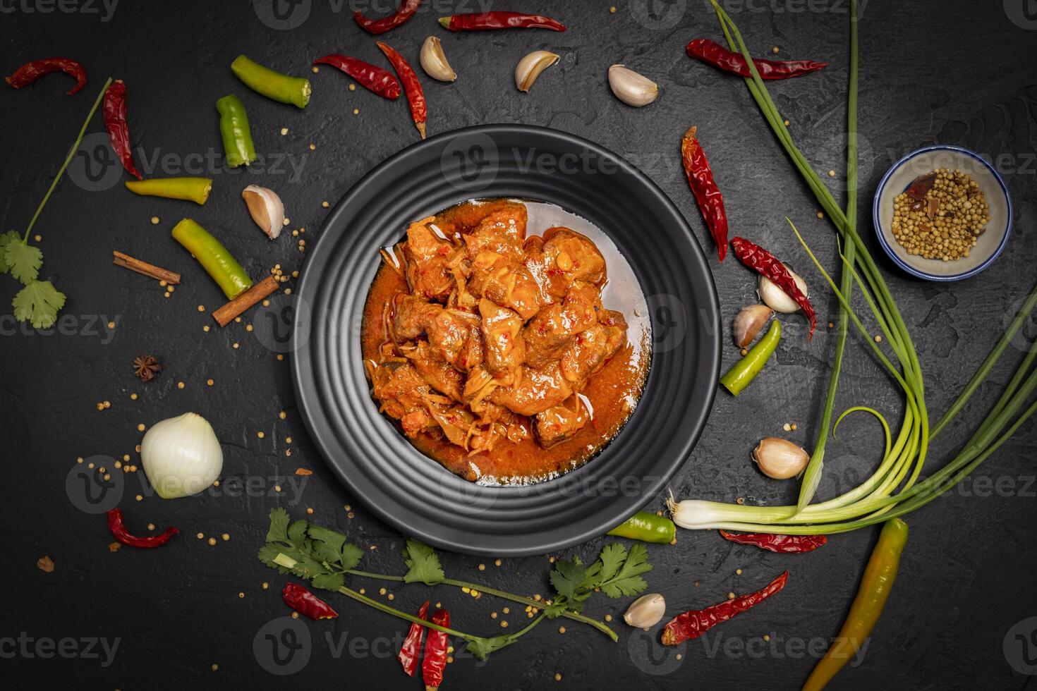 Delicious beef or pork stew in a rustic black plate with fresh coriander, red chilies, garlic, onions and spices placed on a black background, top view photo