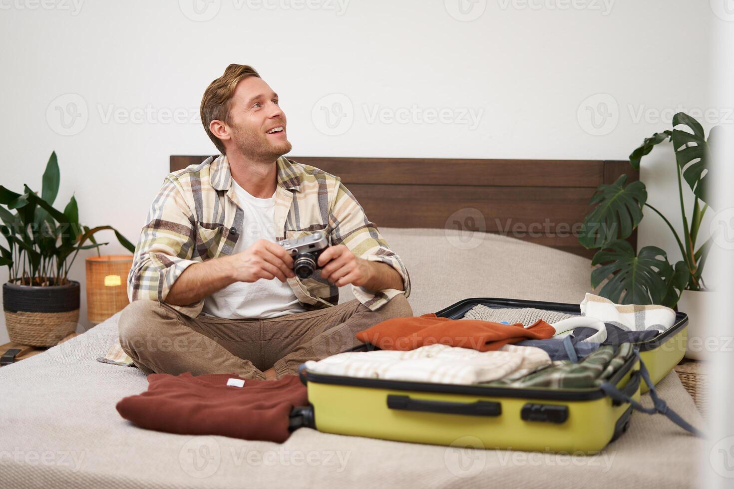 retrato de feliz, sonriente joven hombre sentado en cama, desempacar su maleta después un día festivo, turista mirando a su foto álbum en cámara