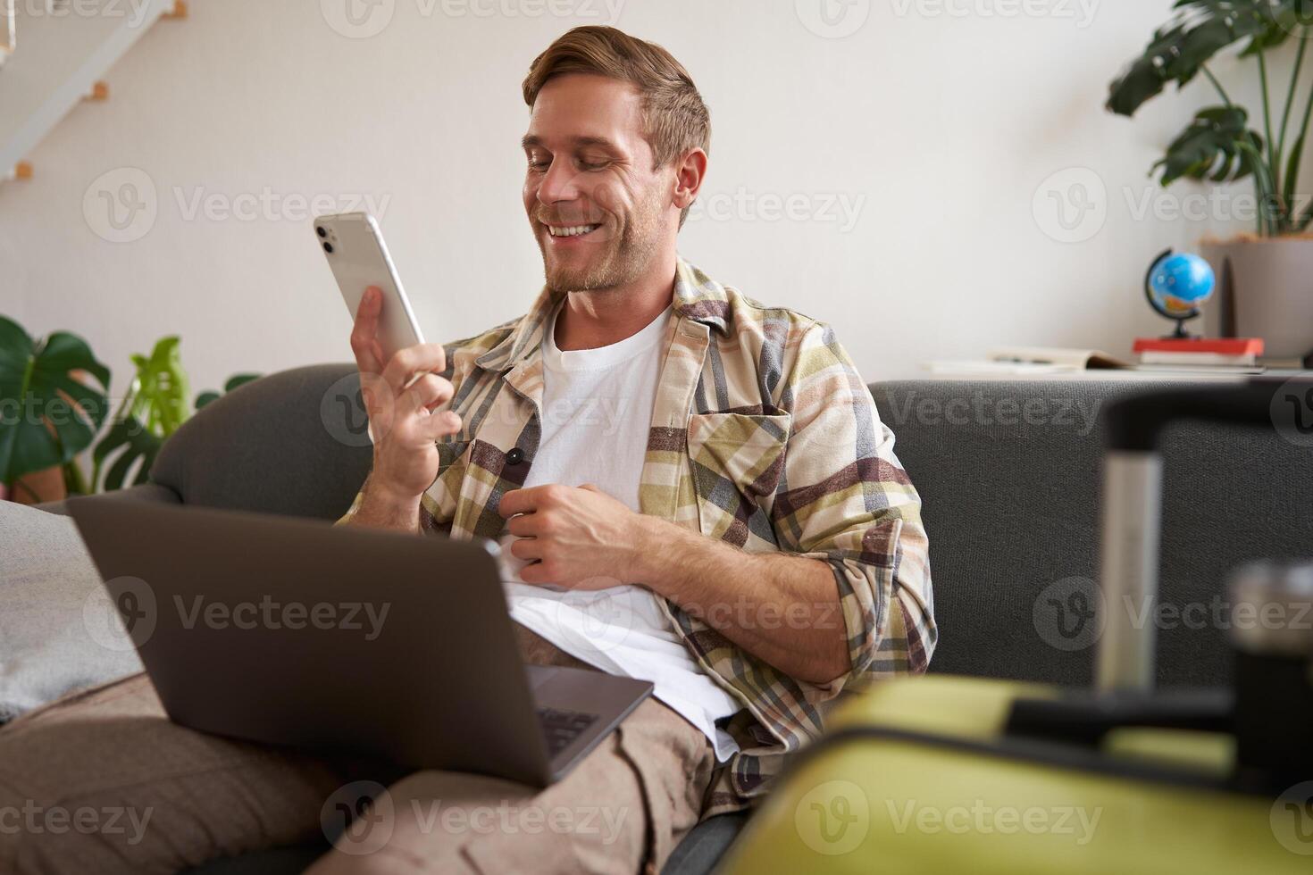 Smiling handsome man going on holiday, making a phone call, holding laptop, prepared suitcase for a trip abroad photo