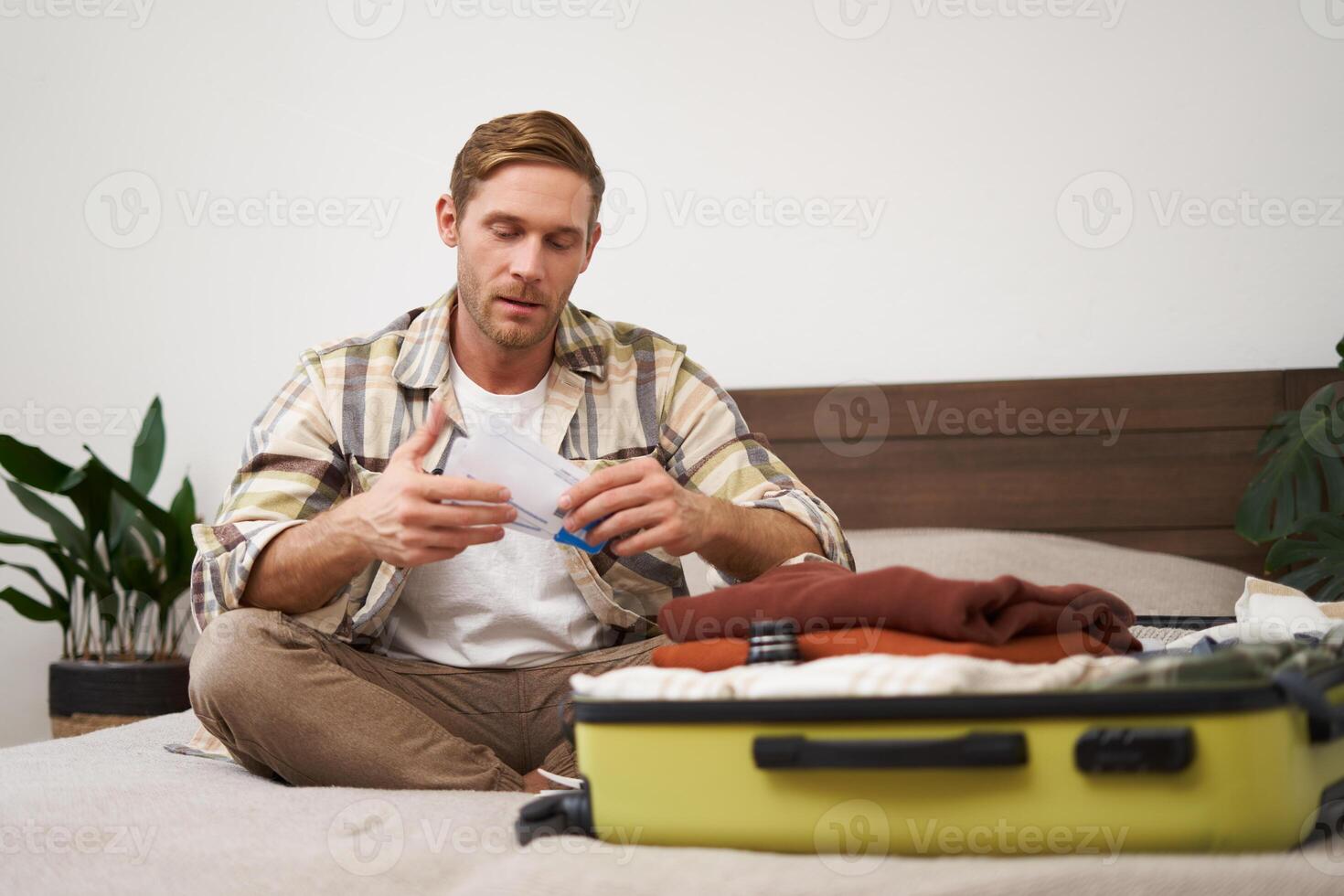 Portrait of handsome man, tourist, looking at his holiday tickets, booked vacation with travel agency, sitting on bed and packing clothes in suitcase photo