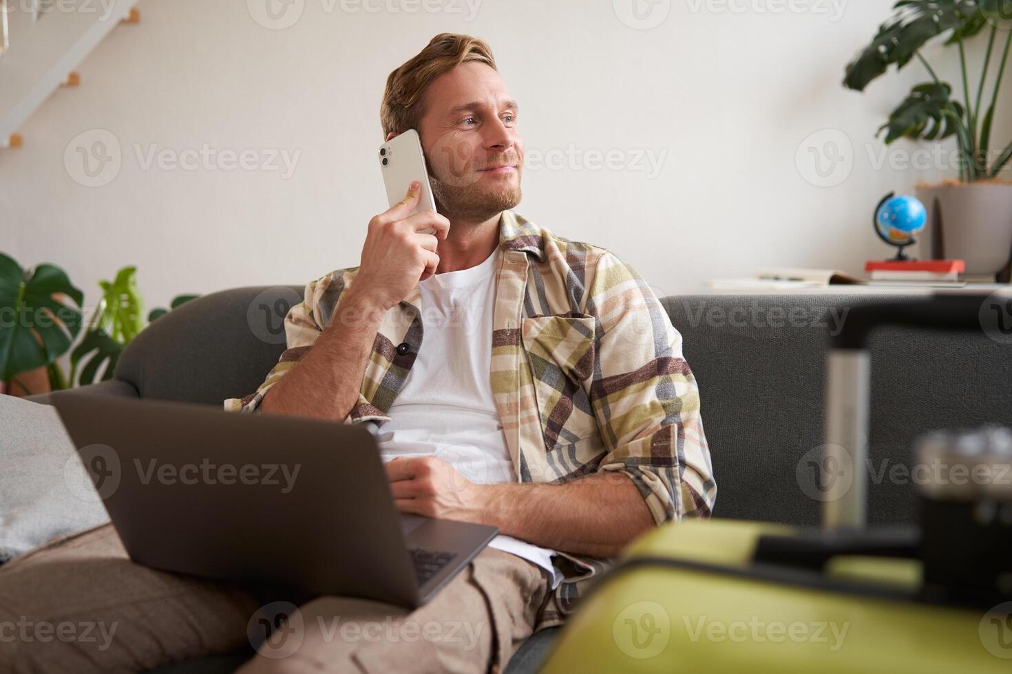 Handsome smiling man with laptop, making a phone call, looking aside while waiting on the telephone, confirms booking online photo