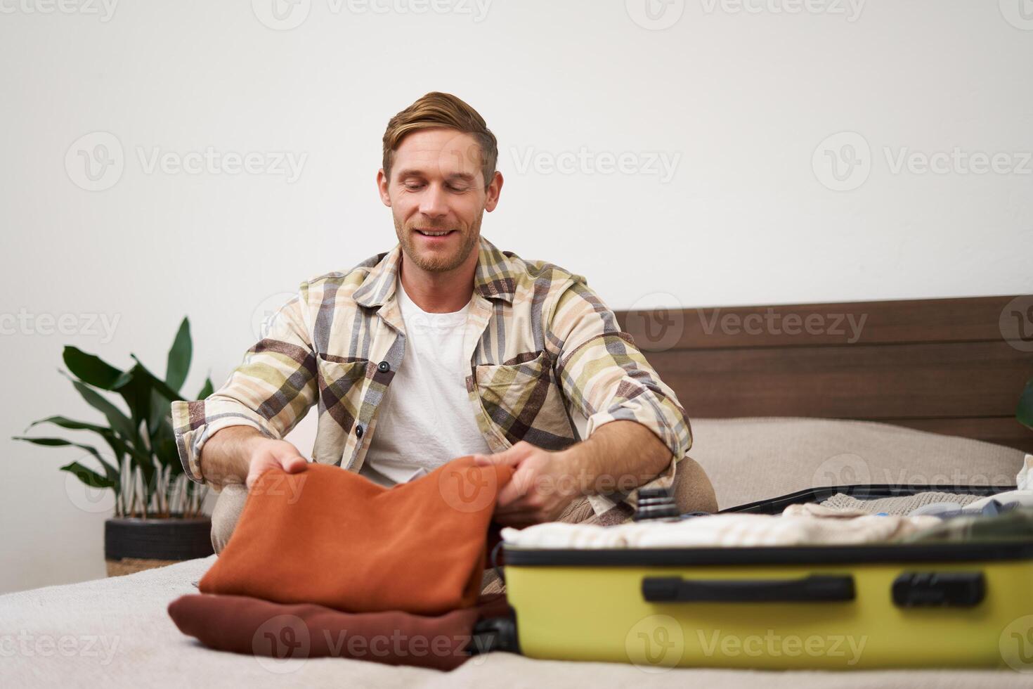 Portrait of handsome young man, tourist unpacking clothes in hotel room, staying in hostel on vacation, travelling, sitting with luggage on bed photo
